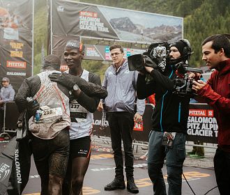 Zwei Athleten umarmen sich am Ziel des Pitz Alpine Glacier Trails, Teil der renommierten Golden Trail World Series, umgeben von Kameraleuten und beeindruckender Kulisse.