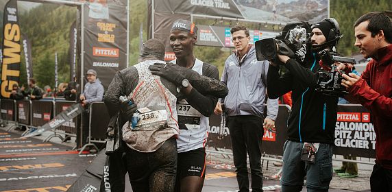 Zwei Athleten umarmen sich am Ziel des Pitz Alpine Glacier Trails, Teil der renommierten Golden Trail World Series, umgeben von Kameraleuten und beeindruckender Kulisse.