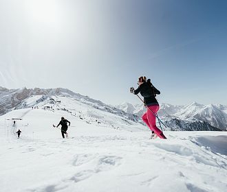 Menschen laufen mit Skistöcken durch eine schneebedeckte Berglandschaft bei klarem Himmel. Im Hintergrund sind beeindruckende Gipfel zu sehen.