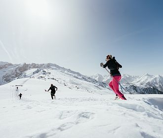 Menschen laufen mit Skistöcken durch eine schneebedeckte Berglandschaft bei klarem Himmel. Im Hintergrund sind beeindruckende Gipfel zu sehen.