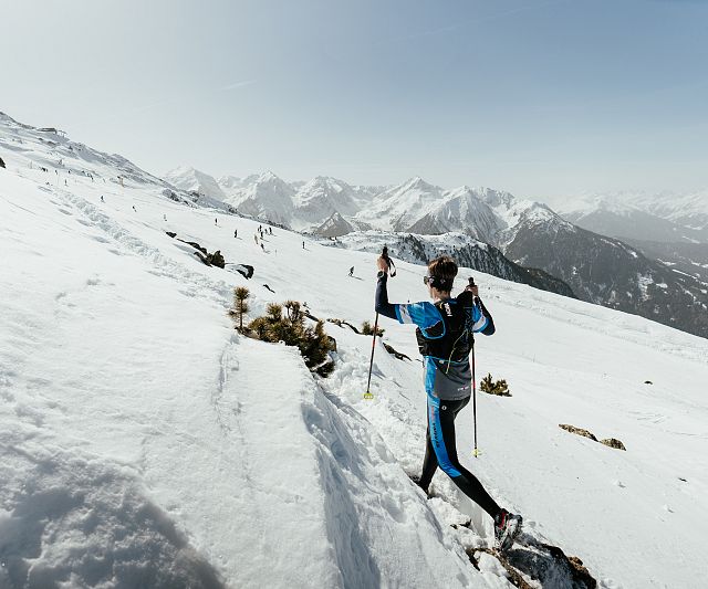 Ein Trailrunner in blauer Kleidung erklimmt einen schneebedeckten Hang in einer bergigen, verschneiten Landschaft unter klarem Himmel, umgeben von majestätischen Gipfeln.