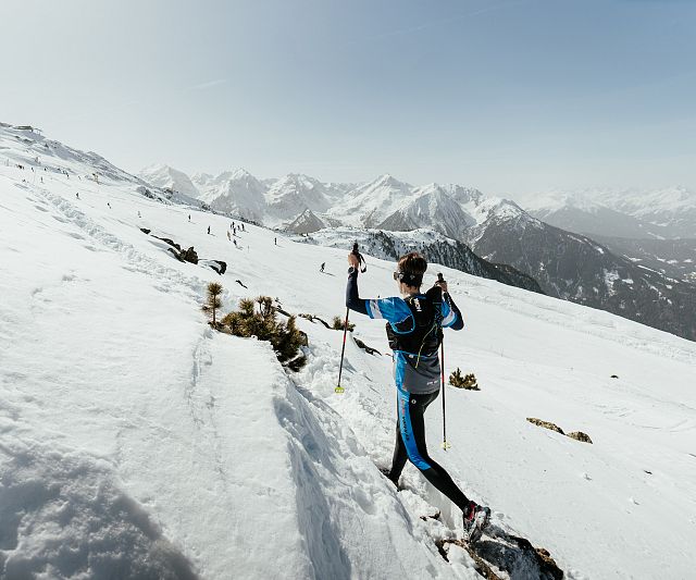 Ein Trailrunner in blauer Kleidung erklimmt einen schneebedeckten Hang in einer bergigen, verschneiten Landschaft unter klarem Himmel, umgeben von majestätischen Gipfeln.