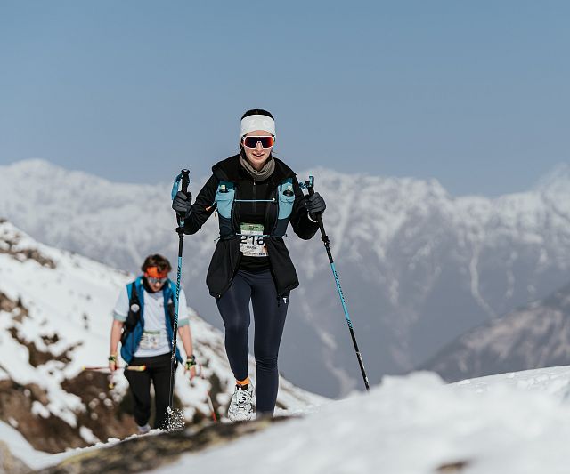 Zwei Sportler wandern in winterlicher Berglandschaft auf einem schneebedeckten Pfad. Sie tragen warme Kleidung, Sonnenbrillen und verwenden Wanderstöcke.