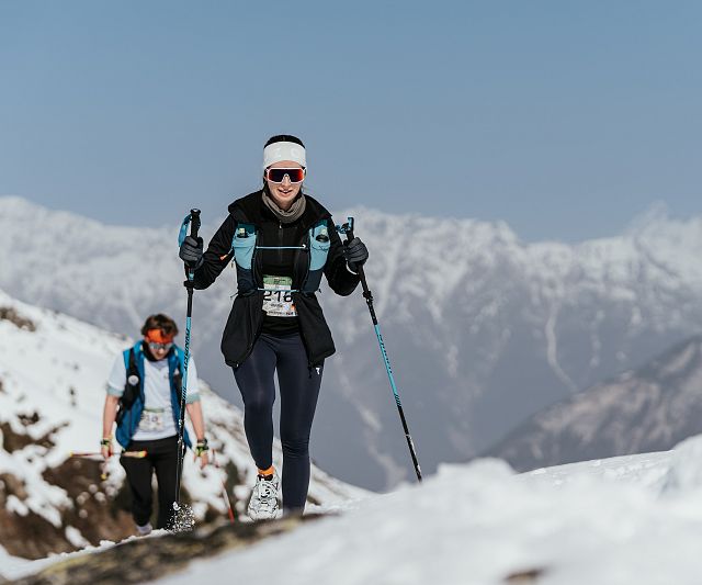 Zwei Sportler wandern in winterlicher Berglandschaft auf einem schneebedeckten Pfad. Sie tragen warme Kleidung, Sonnenbrillen und verwenden Wanderstöcke.