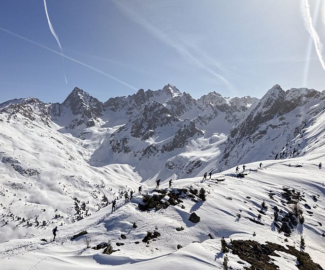 Verschneite Berge unter klarem, blauem Himmel mit Wanderern auf einem Kamm. Sonnenlicht wirft Schatten, während Kondensstreifen den Himmel durchziehen.
