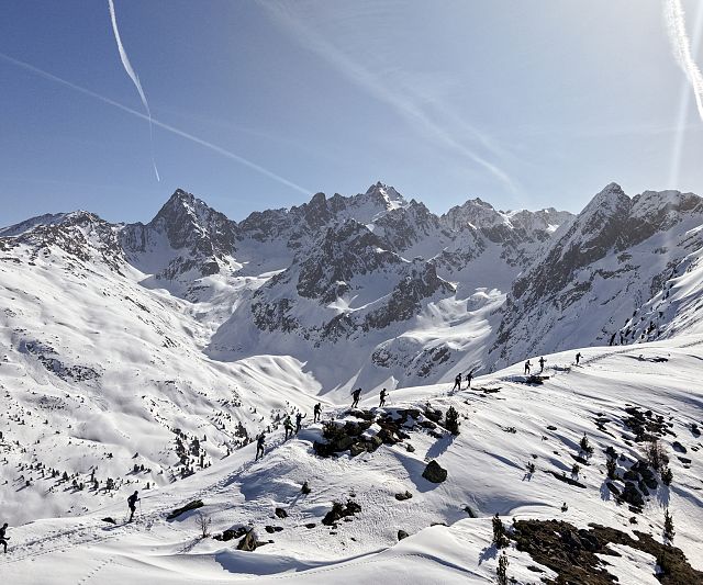 Verschneite Berge unter klarem, blauem Himmel mit Wanderern auf einem Kamm. Sonnenlicht wirft Schatten, während Kondensstreifen den Himmel durchziehen.