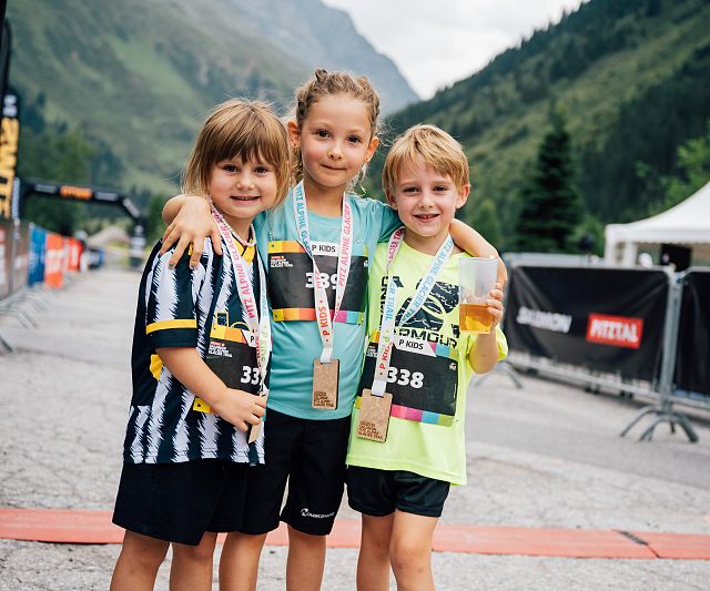 Drei Kinder in Sportkleidung stehen an einer Ziellinie in einer bergigen Landschaft. Sie tragen Medaillen um den Hals und lächeln in die Kamera, zufrieden mit ihrer Leistung.