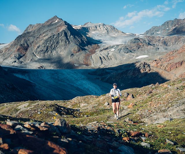 Ein Trailrunner läuft auf einem bergigen Pfad, umgeben von beeindruckenden Gipfeln und Gletschern unter klarem Himmel. Die Landschaft ist steinig und weitläufig.