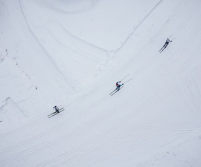 Luftaufnahme von drei Skifahrern auf einer schneebedeckten Piste. Die weiße Schneefläche erstreckt sich weit, während die Spuren der Skier klare Linien bilden.