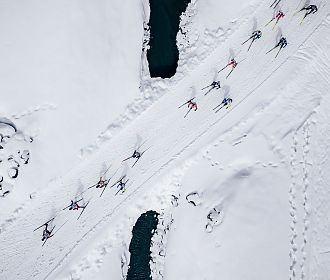 Luftaufnahme von Skilangläufern, die eine schneebedeckte Landschaft überqueren. Die Spuren ihrer Bewegungen sind im Schnee sichtbar, umgeben von tiefen, eisigen Gewässern.