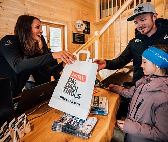 In einem Holzgebäude überreicht eine Frau einem Mann eine Papiertüte mit der Aufschrift &quot;Das Dach Tirols&quot;. Ein Kind steht daneben, umgeben von Prospekten und Broschüren.