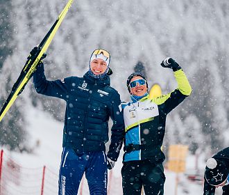 Zwei Personen in Winterkleidung stehen im Schnee. Eine hält Ski und hebt die Hand, die andere zeigt eine Faust. Verschneiter Hintergrund mit Bäumen. Fröhliche Stimmung.