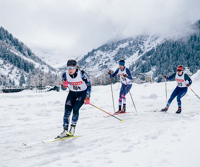 Langläufer beim Rennen durch eine verschneite Berglandschaft. Die Athleten tragen Startnummern und Wintersportkleidung. Im Hintergrund sind bewaldete Berge.