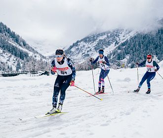 Langläufer beim Rennen durch eine verschneite Berglandschaft. Die Athleten tragen Startnummern und Wintersportkleidung. Im Hintergrund sind bewaldete Berge.