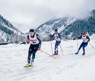 Langläufer beim Rennen durch eine verschneite Berglandschaft. Die Athleten tragen Startnummern und Wintersportkleidung. Im Hintergrund sind bewaldete Berge.