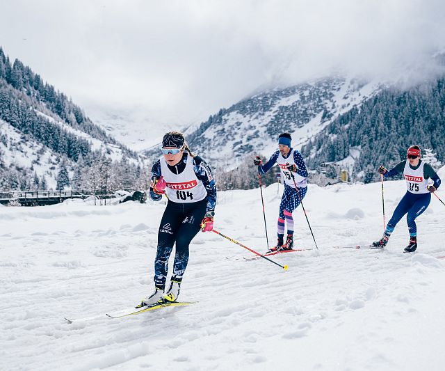 Langläufer beim Rennen durch eine verschneite Berglandschaft. Die Athleten tragen Startnummern und Wintersportkleidung. Im Hintergrund sind bewaldete Berge.
