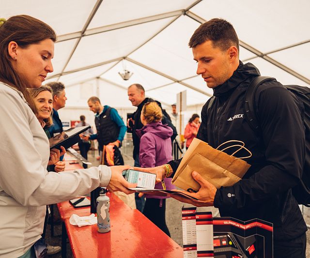 Ein Mann in dunkler Kleidung steht an einem Tisch in einem Zelt und erhält eine Tüte von einer Frau. Mehrere Personen sind im Hintergrund beschäftigt.
