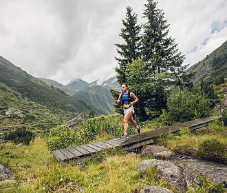 Mädchen beim Trailrunning überquert einen Holzsteg in einer grünen, bergigen Landschaft mit Tannen im Hintergrund.