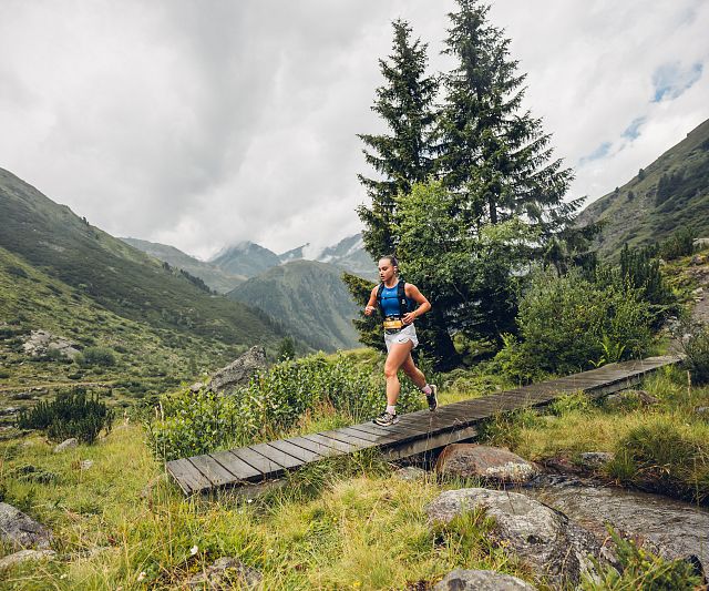 Mädchen beim Trailrunning überquert einen Holzsteg in einer grünen, bergigen Landschaft mit Tannen im Hintergrund.