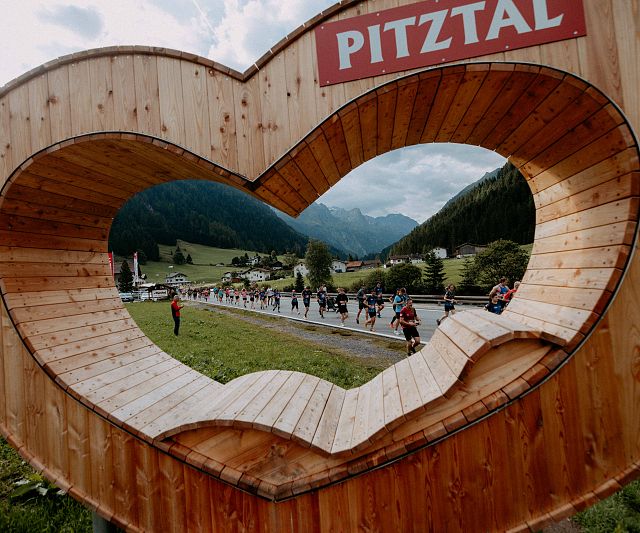 Ein großes hölzernes Herz mit der Aufschrift &quot;Pitztal&quot; bietet einen Rahmen für die Aussicht auf ein malerisches Tal mit Bergen und Läuferinnen und Läufer des Gletschermarathons.