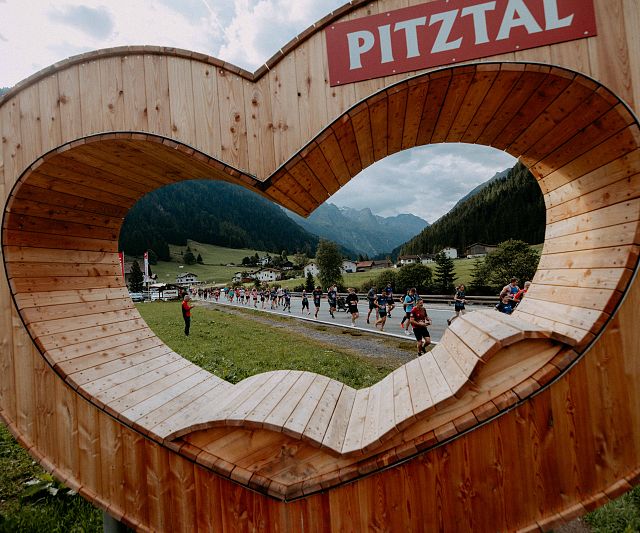 Ein großes hölzernes Herz mit der Aufschrift &quot;Pitztal&quot; bietet einen Rahmen für die Aussicht auf ein malerisches Tal mit Bergen und Läuferinnen und Läufer des Gletschermarathons.