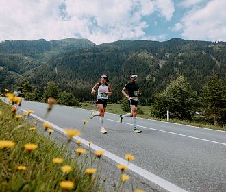 Zwei Personen joggen auf einer Landstraße mit blühenden Wiesen und bewaldeten Bergen im Hintergrund. Der Himmel ist teilweise bewölkt. Eine friedliche Szene.
