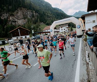 Läuferinnen und Läufer starten bei einem Marathon in einer malerischen Berglandschaft. Zuschauer feuern sie von der Seite an. Startbogen und alpine Kulisse sind sichtbar.