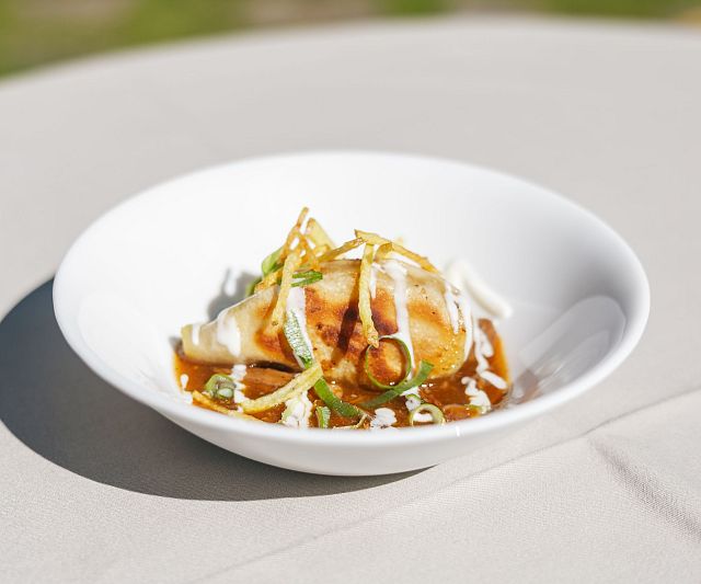 Ein Gericht auf einem weißen Teller, das eine gefüllte Teigtasche zeigt, garniert mit einer Sauce, Zwiebelstreifen und Kräutern. Der Hintergrund ist unscharf.