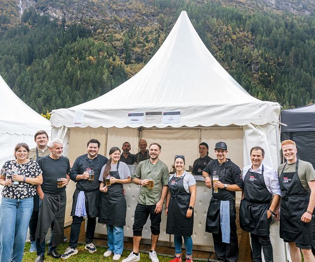 Eine Gruppe von Menschen steht vor einem weißen Zelt auf einem Festival. Sie tragen Schürzen und halten Getränke, umgeben von einer malerischen Berglandschaft.