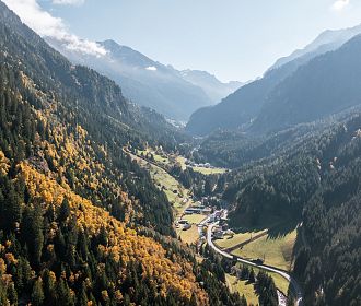 Luftaufnahme eines malerischen Tals in den Alpen, umgeben von grünen und herbstlich gefärbten Wäldern. Eine kleine Ortschaft und Straßen schlängeln sich durch die Landschaft.