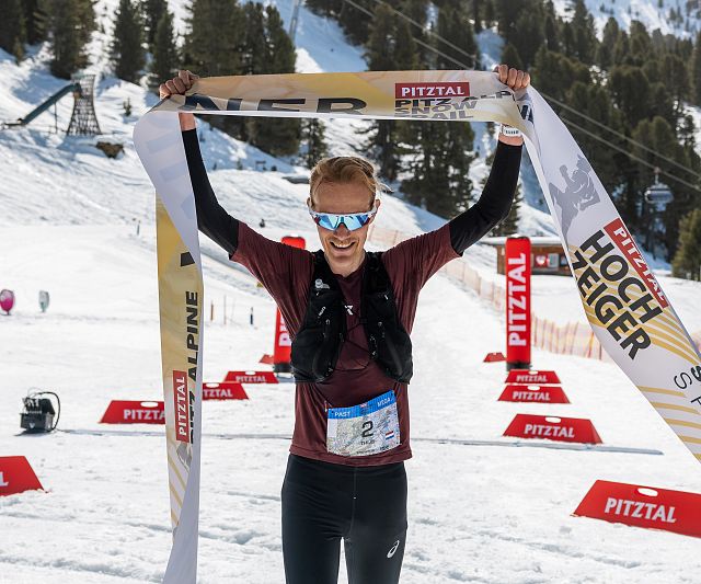 Ein Sportler jubelt beim Überqueren der Ziellinie in einer Schneelandschaft. Er trägt Sonnenbrillen und hebt das Siegerband. Im Hintergrund sind Bäume und Berge sichtbar.