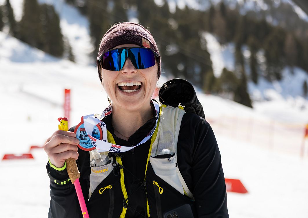 Frau in Sportkleidung steht lächelnd im Schnee vor einem Ski-Hang, hält eine Medaille und einen Skistock. Sie trägt eine Sonnenbrille und eine warme Mütze.