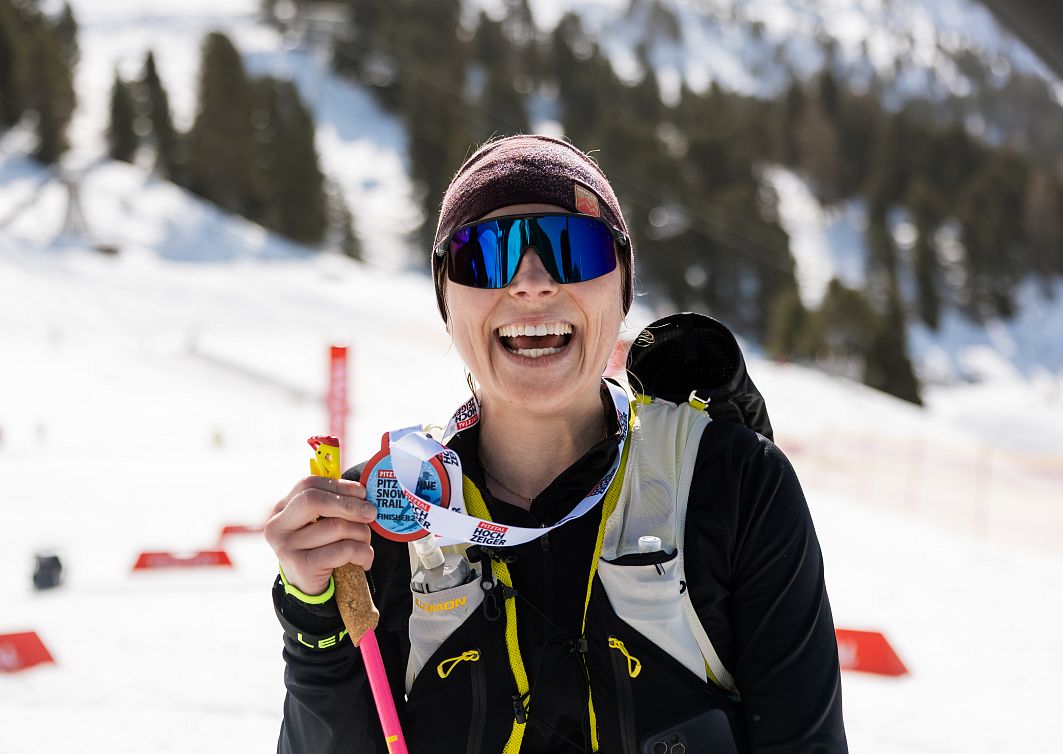 Frau in Sportkleidung steht lächelnd im Schnee vor einem Ski-Hang, hält eine Medaille und einen Skistock. Sie trägt eine Sonnenbrille und eine warme Mütze.