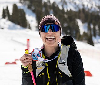 Frau in Sportkleidung steht lächelnd im Schnee vor einem Ski-Hang, hält eine Medaille und einen Skistock. Sie trägt eine Sonnenbrille und eine warme Mütze.
