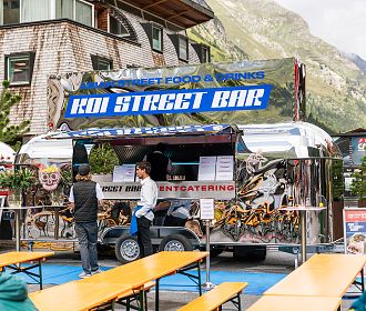 Ein Lebensmittelstand mit der Aufschrift "Koi Street Bar". Der Stand ist silber und hat bunte Grafiken. Zwei Personen stehen davor und eine Tafel zeigt das Menü an.