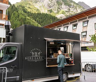Ein schwarzer Food Truck mit der Aufschrift 'Berggams' steht in einer ländlichen Umgebung. Im Hintergrund sind Berge und ein Gebäude zu sehen, während eine Person am Truck steht.