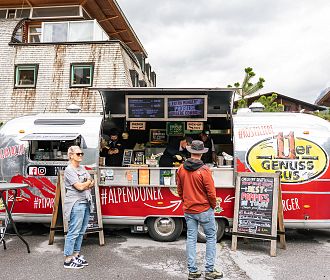 Ein Food Truck im Retro-Stil in einer urbanen Umgebung, mit Menütafeln, Kunden, die bestellen, und einer einladenden Atmosphäre. Wetter ist bewölkt.