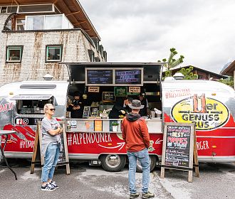 Ein Foodtruck im Retro-Stil in einem urbanen Umfeld. Zwei Personen stehen vor dem Truck, während der Verkäufer im Inneren die Speisekarte anzeigt. Wolkiger Himmel.