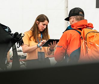 Eine Frau in einem orangefarbenen Shirt nutzt ein Tablet, während sie mit einem Mann in orangefarbener Jacke und Rucksack spricht. Die Szene wirkt geschäftlich und konzentriert.