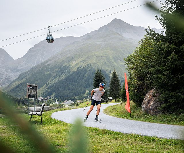 Ein Ski-Roller Athletin fährt auf einem geschwungenen Weg durch eine grüne Berglandschaft. Im Hintergrund sind Berge und die Rifflsee Seilbahn zu sehen. Bäume säumen den Weg.