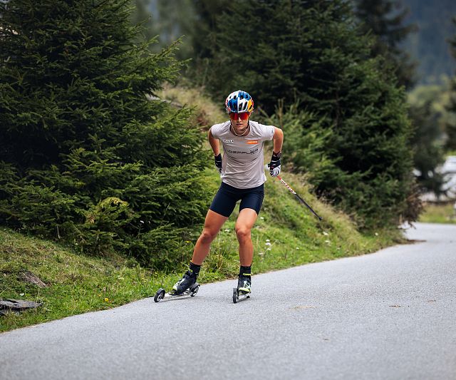 Eine Athletin mit Helm und Sportsachen fährt auf einer kurvigen, asphaltierten Straße durch eine bewaldete Landschaft auf Skiroller. Die Szene wirkt sportlich und dynamisch.