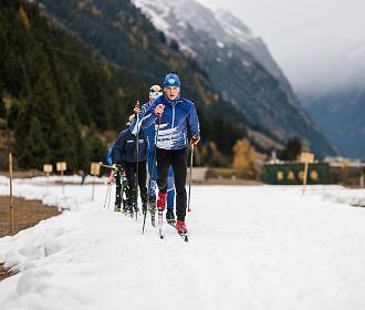 Eine Gruppe von Langläufern in blauer Kleidung gleitet über einen schneebedeckten Weg. Umgeben von Bergen und Nadelbäumen, unter bewölktem Himmel.