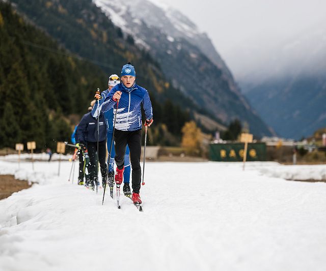 Eine Gruppe von Langläufern in blauer Kleidung gleitet über einen schneebedeckten Weg. Umgeben von Bergen und Nadelbäumen, unter bewölktem Himmel.