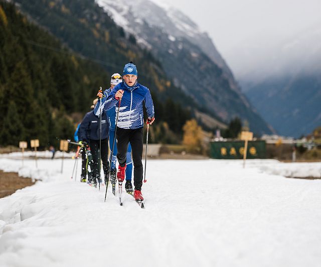 Eine Gruppe von vier Langläufern auf einer schneebedeckten Strecke in den Bergen. Sie tragen sportliche Bekleidung und Ski-Ausrüstung, während im Hintergrund die Landschaft zu sehen ist.
