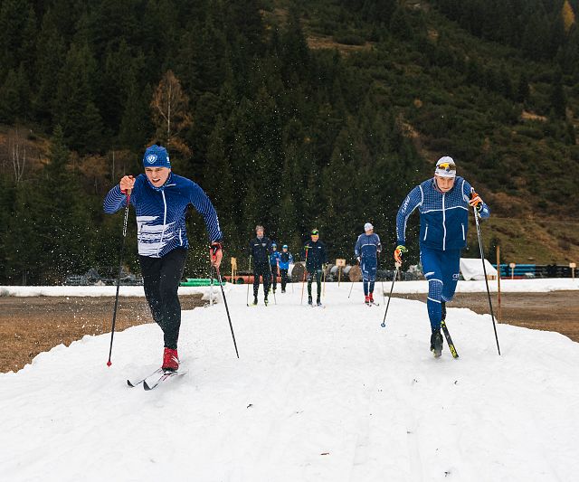 Zwei Langläufer in blauen Anzügen und Helmen gleiten dynamisch über eine schneebedeckte Strecke. Im Hintergrund sind Bäume und mehrere Menschen erkennbar.