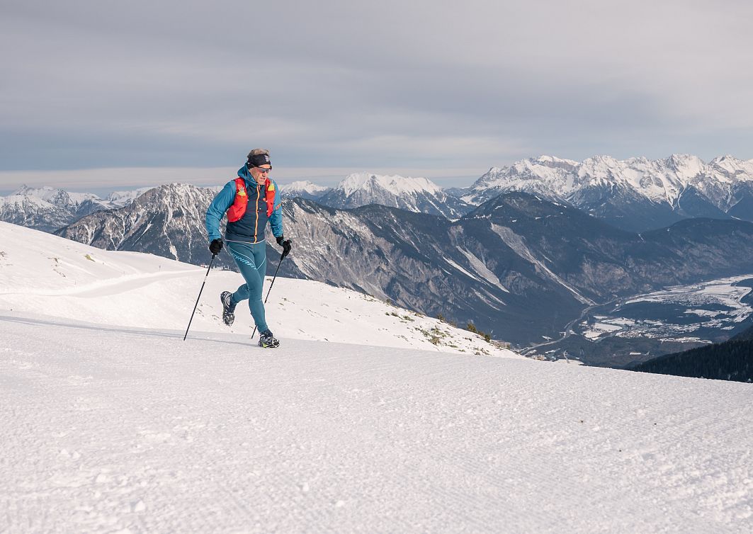 Ein Trilrunner läuft in Winterkleidung über schneebedeckte Berge. Der Himmel ist bewölkt, und im Hintergrund sind beeindruckende Bergketten zu sehen.