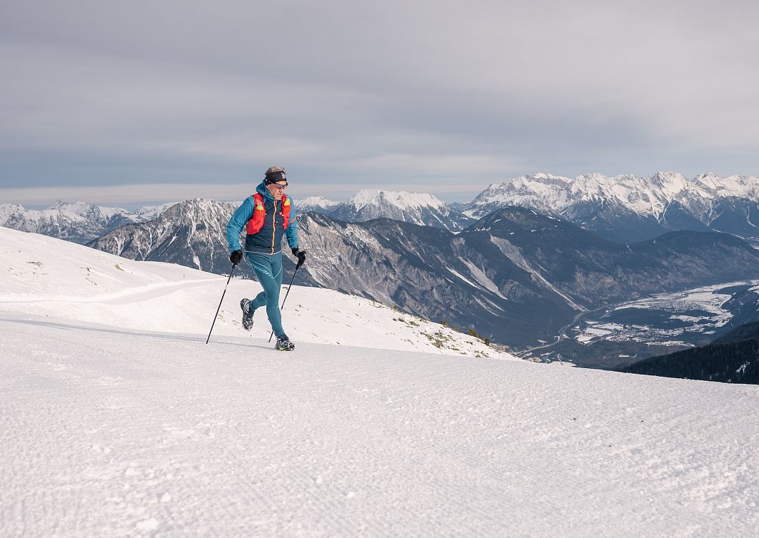 Ein Trilrunner läuft in Winterkleidung über schneebedeckte Berge. Der Himmel ist bewölkt, und im Hintergrund sind beeindruckende Bergketten zu sehen.