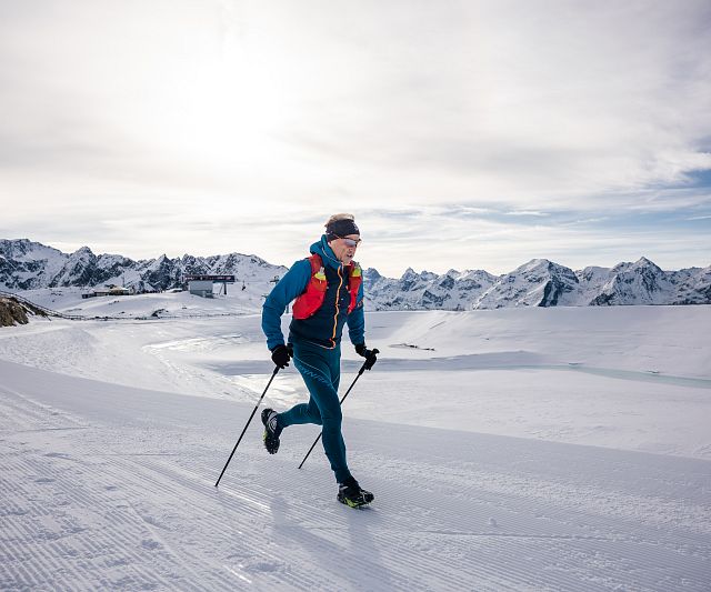 Eine Trailrunner im blauen Anzug läuft mit Skistöcken über schneebedeckte Berge. Der Himmel ist bewölkt, und im Hintergrund sind beeindruckende Berggipfel zu sehen.