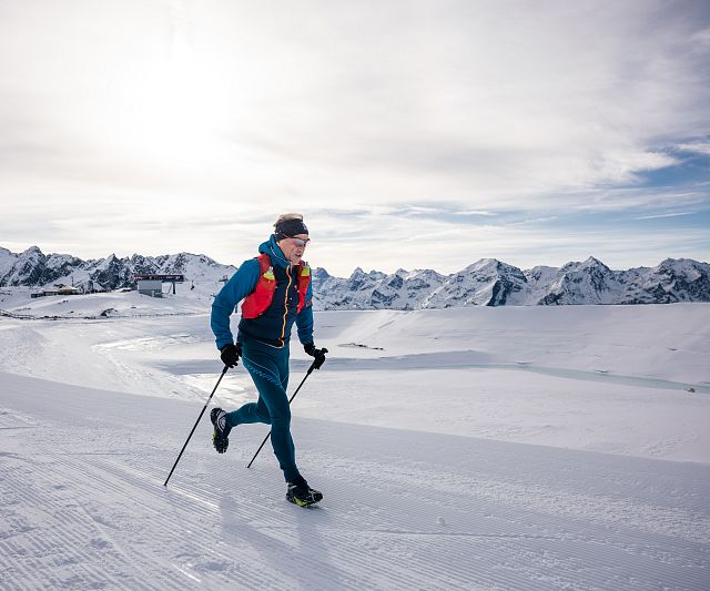 Eine Trailrunner im blauen Anzug läuft mit Skistöcken über schneebedeckte Berge. Der Himmel ist bewölkt, und im Hintergrund sind beeindruckende Berggipfel zu sehen.