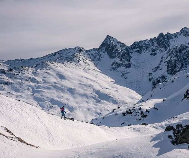 Eine einzelne Person wandert durch eine verschneite Berglandschaft mit markanten Gipfeln im Hintergrund. Der Himmel ist leicht bewölkt und die Stimmung wirkt ruhig und still.
